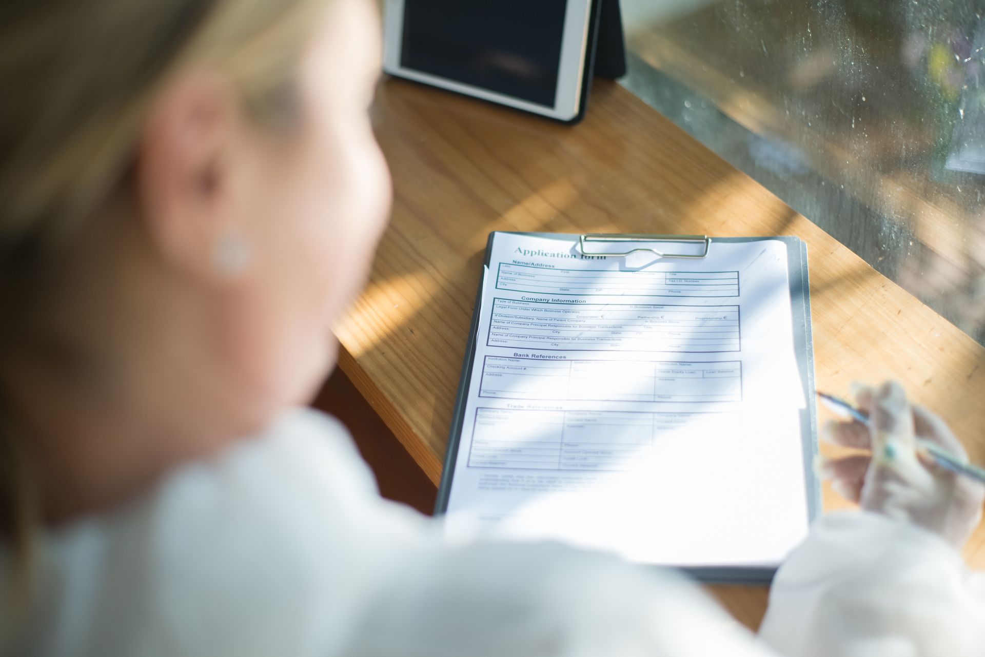 A woman fills out an application form with a pen at a sunlit wooden desk