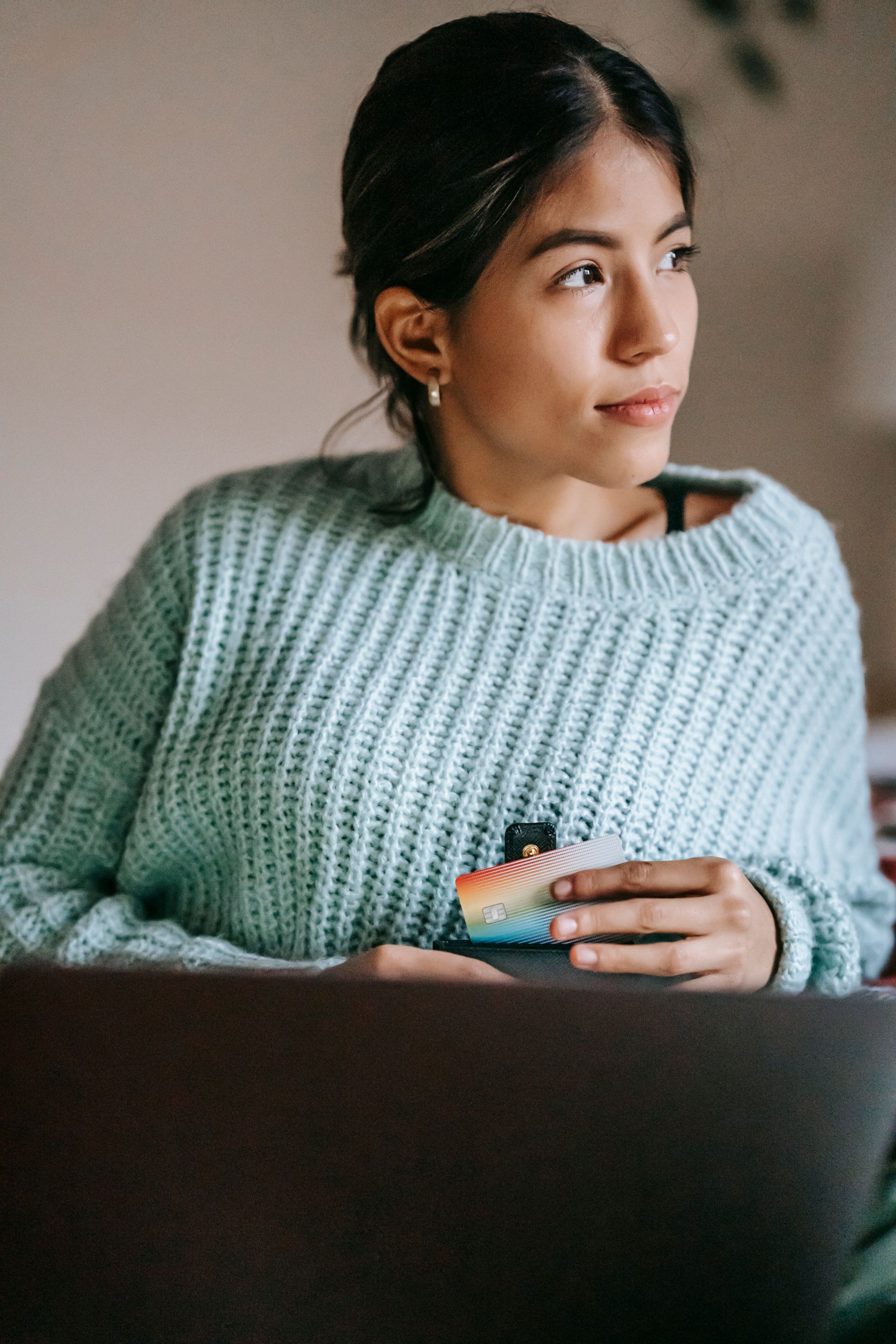 Young woman in a cozy sweater holding a credit card, contemplating an online purchase