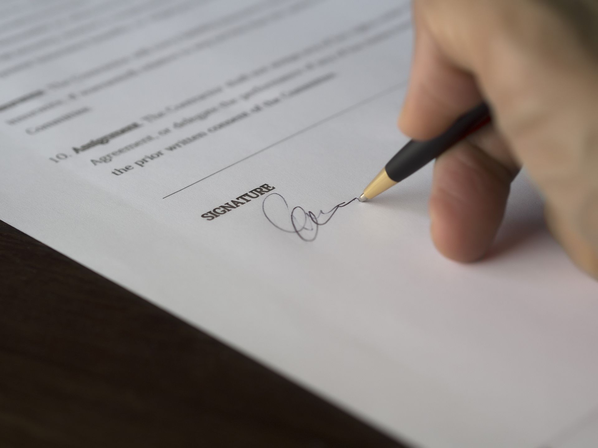 A hand signs a formal contract with a pen on a wooden desk