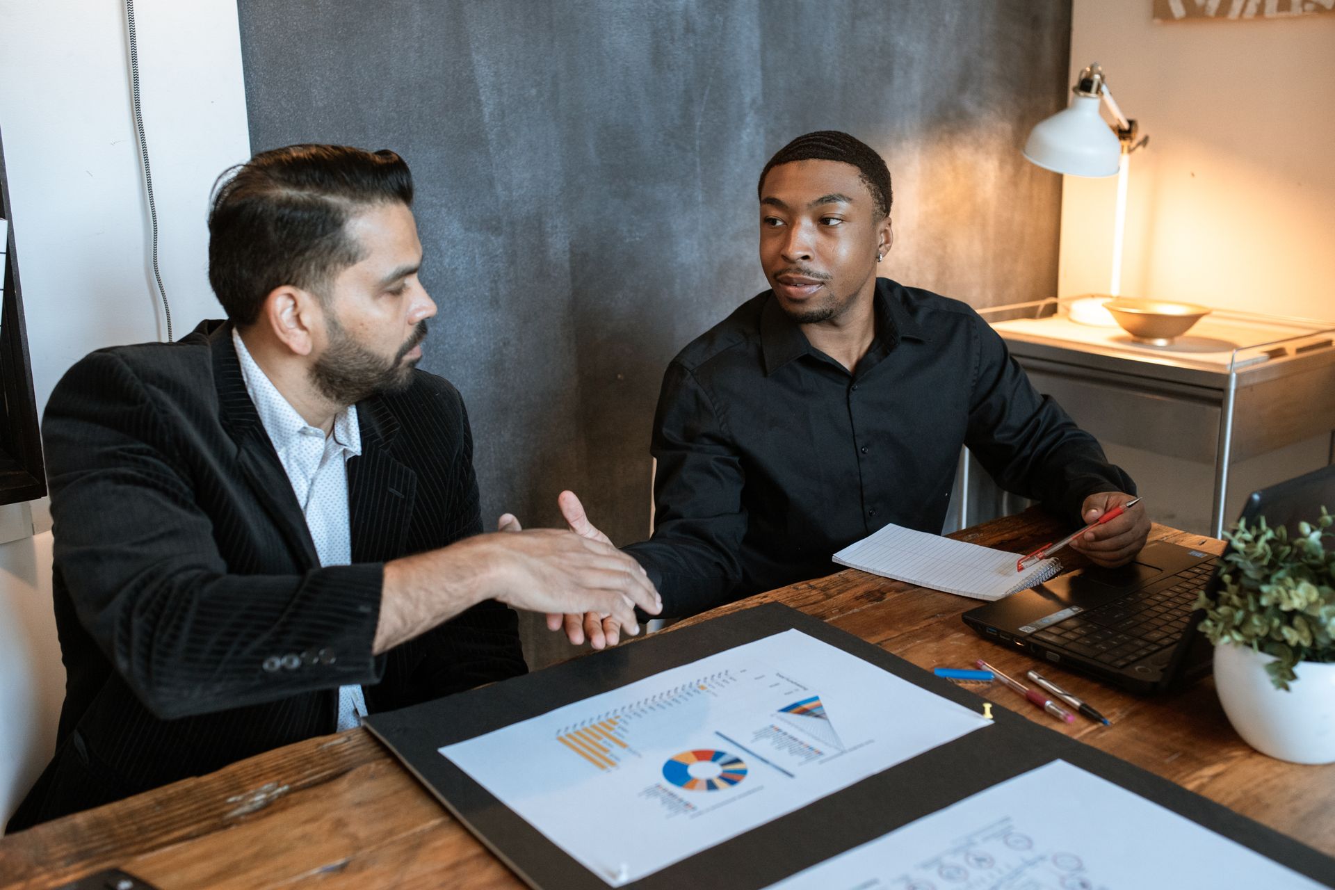 Two businessmen shaking hands over a desk with charts in a modern office